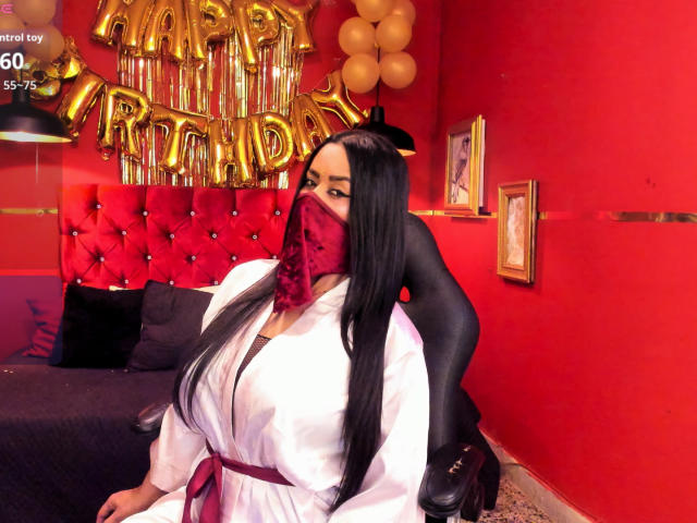 AnissaAli with long black hair, wearing a white robe, red mask, and black stockings, sitting on a bed with a red tufted headboard, her tits slightly visible.