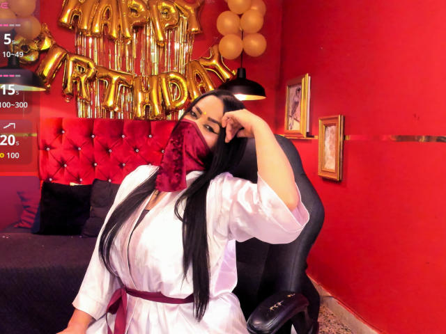 Anissa Ali mit langem, schwarzem Haar, die ein weißes Gewand und eine rote Maske trägt, sitzt auf einem schwarzen Stuhl, berührt ihre Stirn und zeigt ihre großen Brüste, in einem roten Raum..