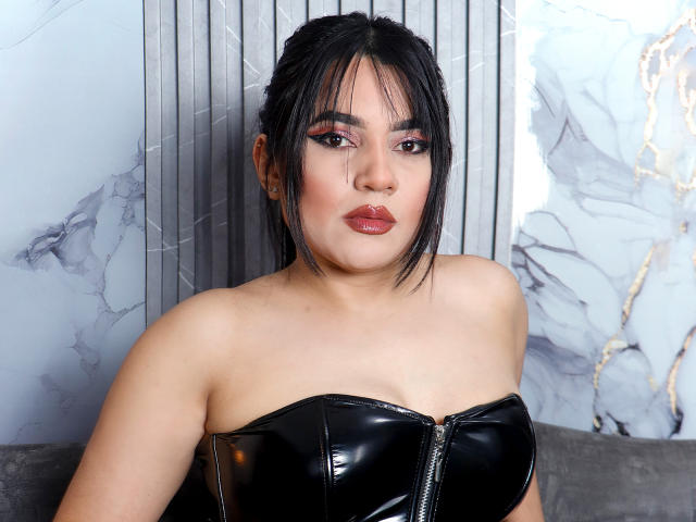 AliceCarusso, com cabelo escuro, batom vermelho e blusa de couro preta sem alças, mostrando os seios, usando um piercing no nariz e olhando para a câmera com uma expressão séria..