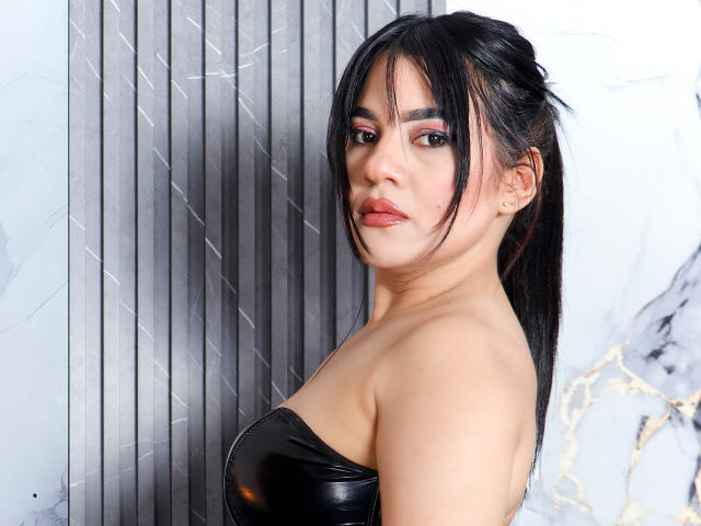 AliceCarusso with black hair in a ponytail, wearing a strapless black leather top, showing her tits, pink eyeshadow, and a slight smile, standing against a gray wall.