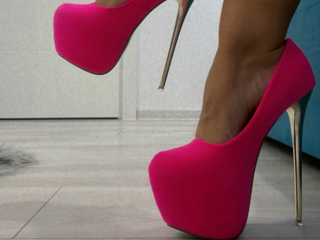 MoniqueBliss's felroze, stilettohakken met zilveren hakken, staan op een grijze houten vloer en laten haar getoonde, bruine benen zien..