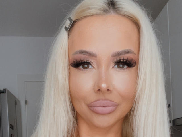 MoniqueBliss has blonde hair, brown eyes, thick long eyelashes, and pinkish-lavender lipstick. She's wearing a small hair clip on the left side of her head.