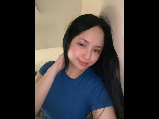 AlaineAtcher, con i capelli neri, indossa una camicia blu, sorride dolcemente, una mano tocca il collo, pelle chiara, trucco leggero, ambiente informale, espressione rilassata..