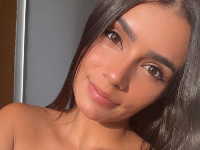 KimmyK has tan skin, brown eyes, and long dark brown hair. She's smiling with pink lips, showing a slight tan line on her shoulders. Her eyebrows are thick and arched.