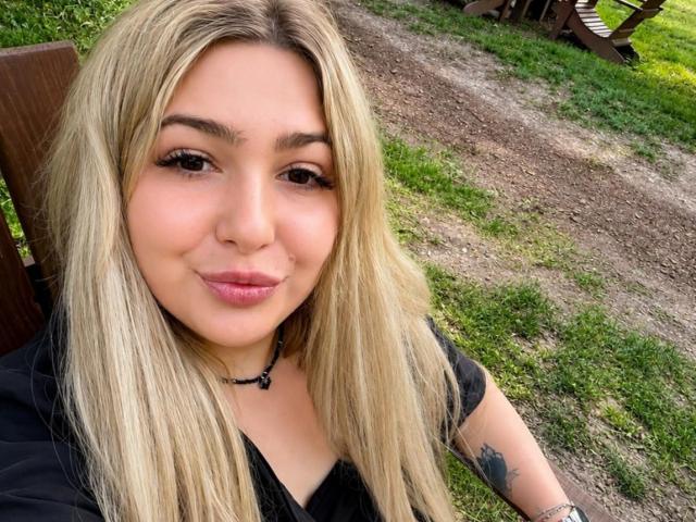 DawnErika with blonde hair, black shirt, and tattoo on right arm, smiling at camera, pink lips, black necklace, green grass and dirt path in background.