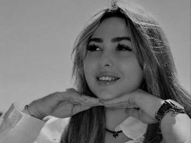 Black-and-white photo of DawnErika with long hair, wearing a white shirt, hands near her face, smiling, visible eyelashes, and a bracelet on her right wrist.