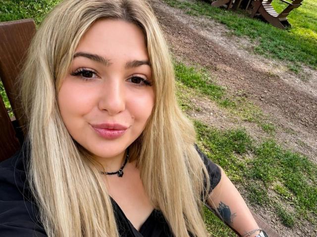 DawnErika with long blonde hair, black shirt, and choker, smiling at the camera, showing a tattoo on her right arm, with pink lips and a cute face.