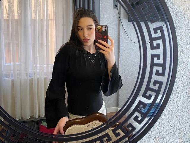 Sherie Stoneback, con cabello largo y castaño, vestida con una camiseta negra de manga larga y pantalones blancos, sentada frente a un espejo circular, sosteniendo un teléfono con un protector de pantalla con motivos griegos..