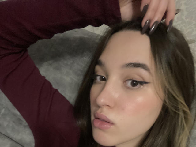Sherie Stoneback, con i capelli castani, le labbra piene e l'eyeliner nero, indossa una camicia a maniche lunghe color borgogna e tocca la testa con le sue unghie lunghe, scure e curate..