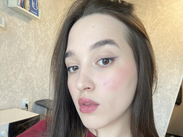 SherieStoneback has pale skin, brown eyes, and pinkish-red lipstick. She has dark brown hair, a small pimple on her nose, and a slightly pouty expression.