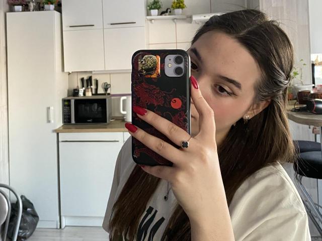SherieStoneback with long brown hair, red nails, holding phone with dragon case, wearing white tee, taking mirror selfie in kitchen, white cabinets in background, black backpack on left.