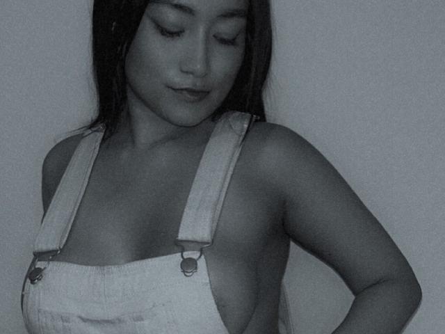 Grayscale photo of VanesaCandy with eyes closed, wearing light denim overalls, exposing her tits, standing against a plain background, slightly smiling, with straight dark hair.