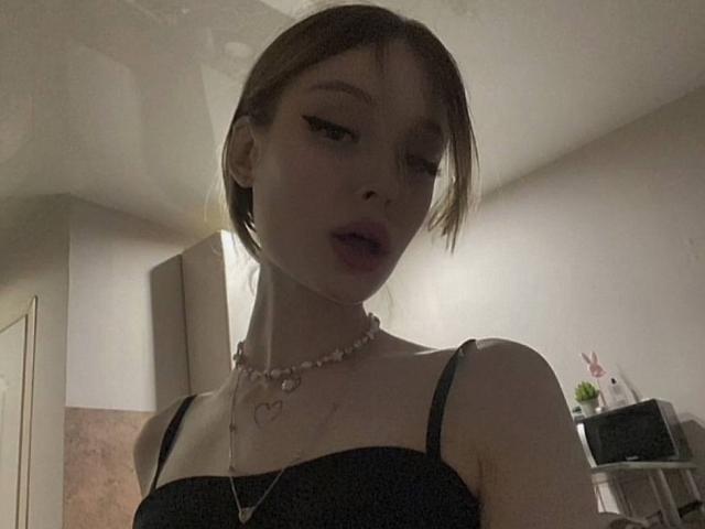 Ceola Goodroe, con cabello castaño corto, un sujetador de encaje negro, un collar rosa y un collar con forma de corazón, labios ligeramente entreabiertos, mirando hacia abajo, iluminación suave, una sombra sutil en el rostro, una habitación en el fondo..