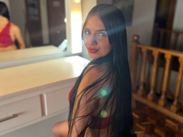 AstraGlow, con lunghi capelli neri, reggiseno rosso e rossetto rosso, guarda oltre la spalla, sorridendo, con un riflesso di luce sulla guancia, seduta vicino a uno specchio. Il seno è visibile..