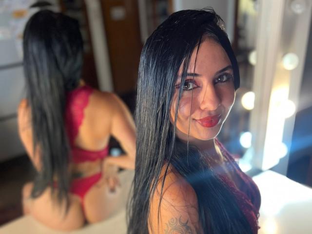 AstraGlow with long black hair, red lingerie, and tattoos, looking back at the camera, showing her ass and smiling, with a mirror reflecting her back.