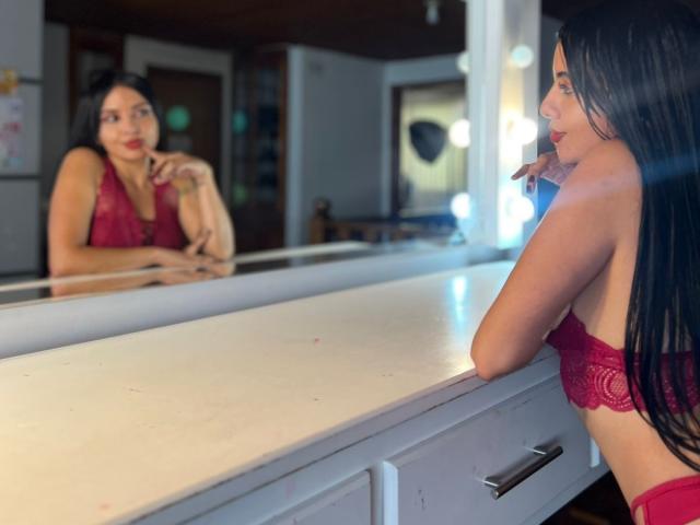 AstraGlow with long black hair, red lace bra, and red panties, leans on a mirror, biting her finger, reflecting her face, showing off her tits and ass.
