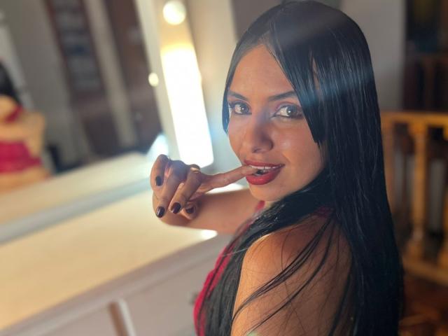 AstraGlow with long black hair, pointing finger at her red-lipsticked mouth, wearing a red and black top, has dark nail polish, and a sexy smile.