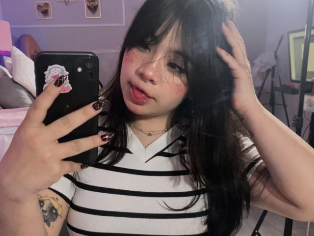 AmaraKazumi, com cabelo preto, língua rosa de fora, sardas, camisa listrada, segurando um telefone, mostrando uma tatuagem no braço esquerdo, com esmalte, selfie no espelho, mãos no cabelo..
