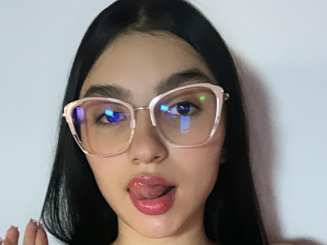 MilanMoon with long black hair, pink glasses reflecting blue and white lights, and her tongue slightly out, showing pink glossy lips. Her hand is partially visible on the left.