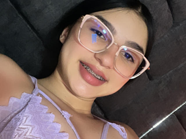 MilanMoon with pink glasses, light purple lace top, smiling, showing her teeth, black hair, cute, reflection in glasses, dark background, soft lighting, looking up.