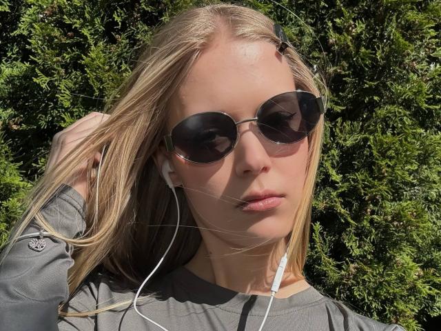 LillianaWhang with blonde hair, grey shirt, black round sunglasses, white earbuds, and slightly parted lips, holding her hair against green bushy background.