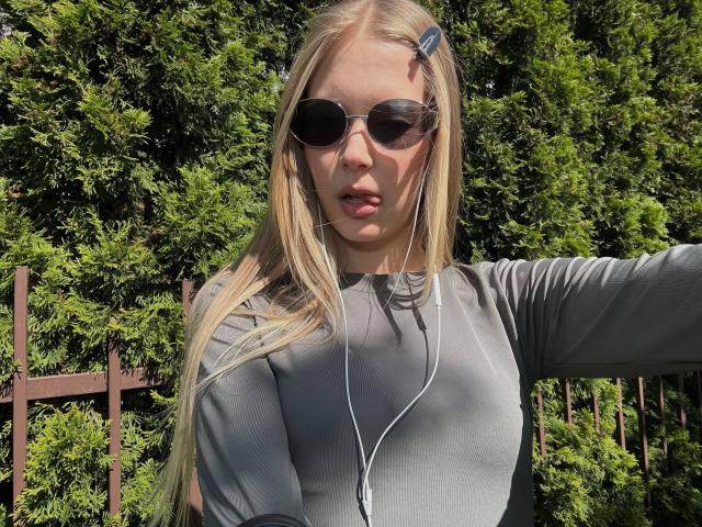 Lilliana Whang, con cabello rubio largo, vestida con una camiseta deportiva gris, gafas de sol negras, audífonos blancos y un clip azul para el cabello, está de pie frente a arbustos verdes. Sus senos son visibles..