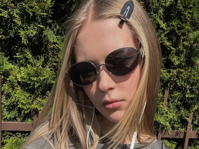 Lilliana Whang mit blonden Haaren, runden schwarzen Sonnenbrillen und einer schwarzen Haarspange, trägt weiße Kopfhörer und wirkt ernst, im Hintergrund ist grünes Laub im Sonnenlicht zu sehen..