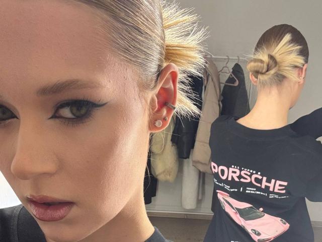 Lilliana Whang mit blondem Dutt, trägt ein schwarzes Hemd, zeigt den Rücken, auf dem "PORSCHE" und ein pinkes Auto abgebildet sind. Sie steht in einem Raum, blickt mit Eyeliner und pinkem Lippenstift in die Kamera..