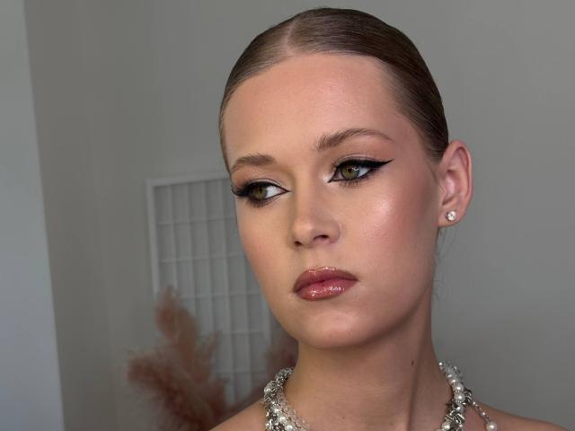 Lilliana Whang ha occhi marroni brillanti, eyeliner nero, labbra rosa lucide e capelli marroni tirati indietro. Indossa una collana di perle e piccoli orecchini di diamanti..