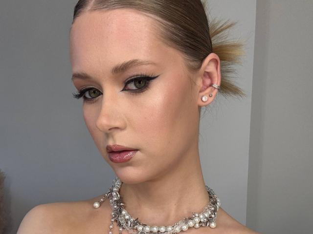 Lilliana Whang heeft glanzende bruine ogen, een vingerhoedje eyeliner en glanzende roze lippen. Ze draagt een parelketting, parel oorbellen en heeft blond haar dat in een strakke knot is opgestoken..