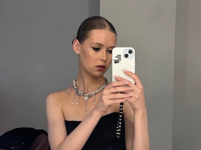 Lilliana Whang, con i capelli castano scuri pettinati all'indietro, un top nero senza spalline, una collana di perle, sta scattando un selfie con un iPhone bianco, con un'espressione seria, pelle chiara, trucco leggero, orecchini visibili..