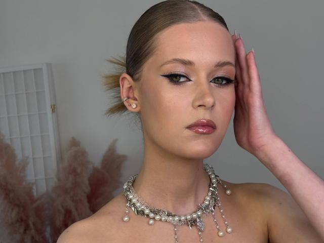 Lilliana Whang ha i capelli biondi tirati indietro, indossa una collana di perle eleganti, con una mano sulla testa, mettendo in risalto un audace eyeliner nero e labbra rosa lucide..