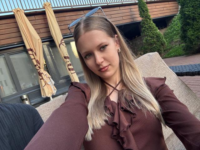 Lilliana Whang heeft blond haar, groene ogen en kleine borsten, en draagt een bruin topje met ruches. Ze staat op een terras met gesloten parasols en een houten gebouw. Ze draagt een zonnebril op haar hoofd..