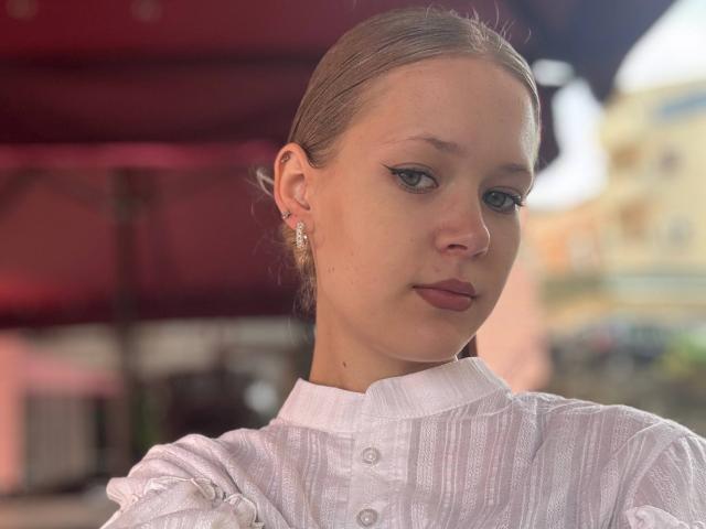 Lilliana Whang tiene la piel clara, ojos verdes y senos pequeños, y lleva una camisa blanca con botones y textura, con mangas abullonadas. Tiene el cabello rubio recogido hacia atrás y está mirando hacia abajo..