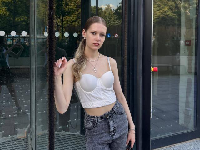 Lilliana Whang, mit kleinen Brüsten, trägt eine weiße, durchsichtige BH-Halter, graue, hoch taillierte Jeans und eine Kreuzkette. Sie steht vor einer Glastür und hält sich ihr Haar..