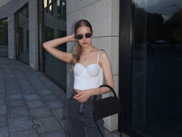 Lilliana Whang, met kleine borsten, draagt een witte bustier, donkere jeans, zwarte zonnebril en een zwarte handtas, en staat voor een glazen gebouw, met haar hand in haar haar, en kijkt naar de camera..