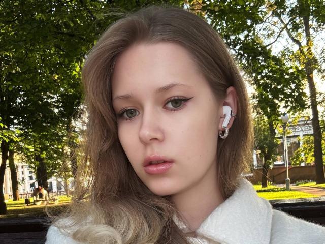 LillianaWhang has pale skin, green eyes, and full pink lips. She's wearing white earbuds, a fluffy white coat, and has long, wavy blonde hair. She looks serious.