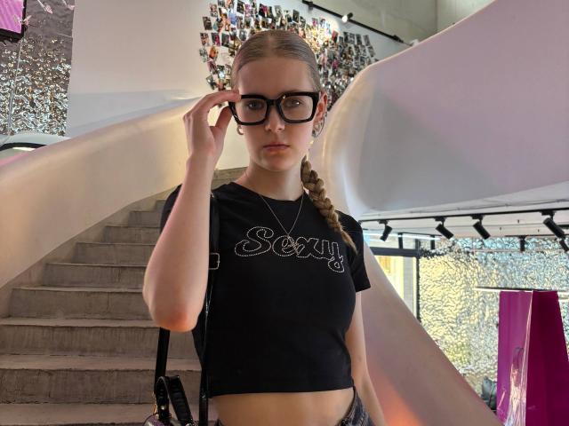 Lilliana Whang, con gafas, top corto negro, trenza, ajustándose las gafas, llevando un collar, de pie frente a una estructura blanca y curva, mostrando un abdomen plano, piel clara, expresión seria..