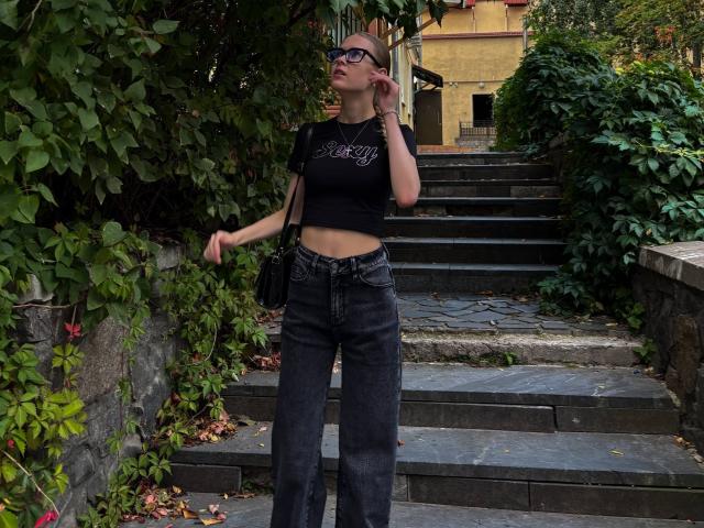 Lilliana Whang è in piedi su dei gradini di pietra, indossa un top corto nero che mette in mostra il seno e dei jeans neri a zampa d'elefante, con una borsa nera. Sullo sfondo si vedono della vegetazione verde e un edificio giallo..