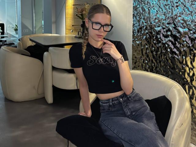 LillianaWhang, with glasses, sitting on a black chair, wearing a black crop top, blue jeans, and black pants, finger near her mouth, blonde braid, medium tits, slim build.