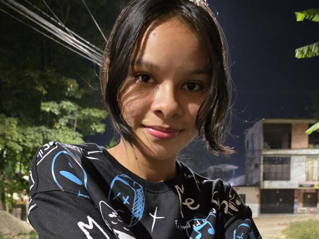 Yurimy with medium brown skin and black hair, wearing a black shirt with blue and white patterns, smiling slightly, showing small pink lips, in a nighttime outdoor setting.