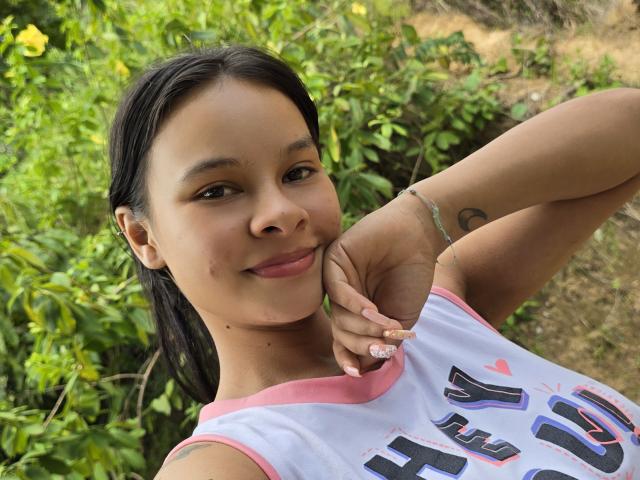 Yurimy with dark hair, brown skin, and small nose, smiling, holding her chin, wearing a pink-trimmed white shirt, showing a tattoo on her wrist, in a green outdoor setting.