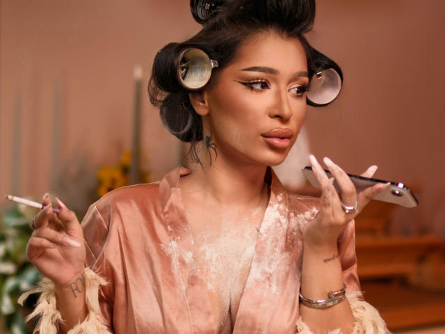 CherrieTresure with curlers in her black hair, wearing a pink, sheer, velvet robe, holds a cigarette in one hand and a mirror in the other, looking at herself.