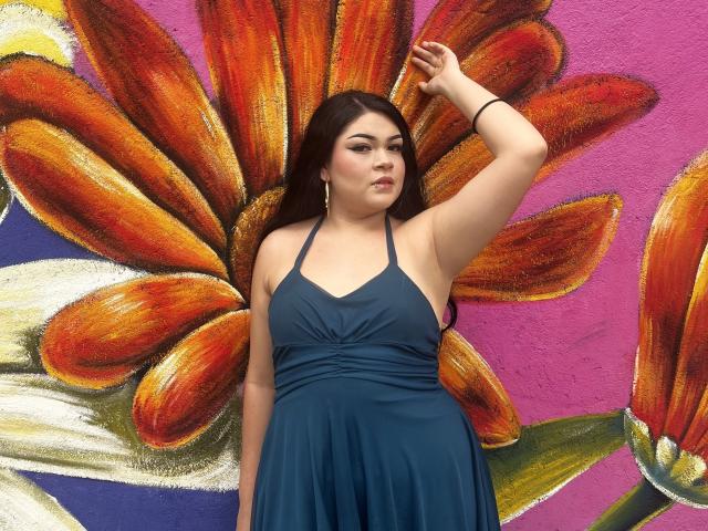 MadissonJoe, com seios grandes, cabelo castanho escuro, vestindo um vestido azul de alças, em pé em frente a uma parede colorida com grandes flores laranjas, seu braço direito levantado, com uma expressão confiante..