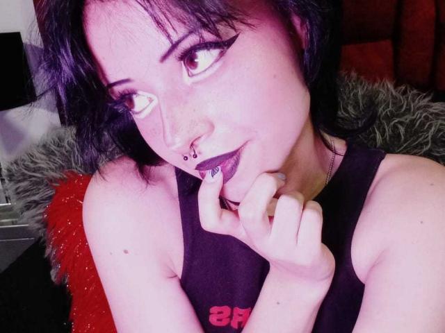 ViolettClay with dark purple lipstick, black sleeveless top, septum piercing, and black eyeliner, biting her finger, looking up with a slight smile, purple-pink lighting.
