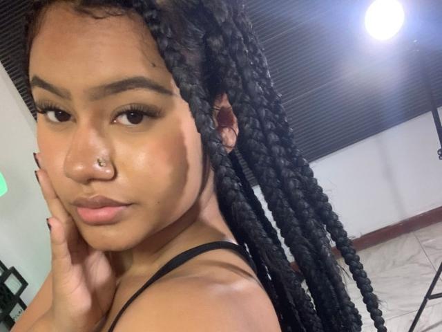 NoiraVale with long black braids, nose ring, and subtle makeup, holding her cheek with a serious expression, wearing a black strap top, under bright studio lights.