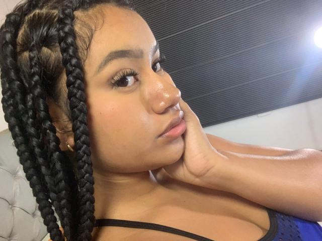 NoiraVale with braided black hair, brown eyes, and glossy skin, resting her chin on her hand, wearing a blue, polka-dot, spaghetti-strap top, looking slightly to the side.