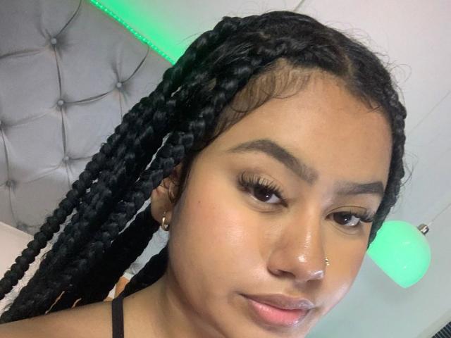 NoiraVale has dark braided hair, a nose piercing, and a neutral expression. She's wearing a black strap, with green LED light in the background.