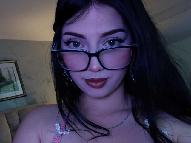 IsabellaaRossi has dark hair, wearing black glasses, red lipstick, and a pink and white striped top, showing off her tits slightly. She's looking at the camera with a smile.