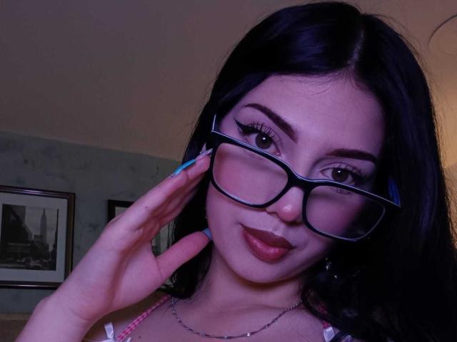 Isabella Rossi, met donker haar, een bril en blauwe nagels, lacht en toont haar hand. Ze draagt een roze top en een ketting, en de foto is genomen bij weinig licht. Haar borsten zijn niet zichtbaar..